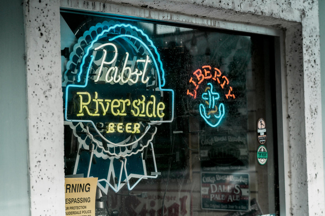 A neon Pabst Riverside Beer sign glowing in a dimly lit bar window, capturing the nostalgic dive bar aesthetic and beer culture vibe.