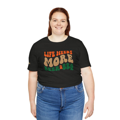 T-shirt with retro-style text reading "Life Needs More Beer & BBQ" in bold, vintage-inspired colors. Perfect for beer lovers, grill masters, and BBQ enthusiasts. Available in plus sizes.