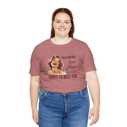 A cheerful, rosy-cheeked pin-up girl with wavy blonde hair smiles brightly. The design features playful retro lettering with the phrase "You smell like fine wine & bad choices – happy to meet you!" in bold, vintage script. Available in plus sizes.
