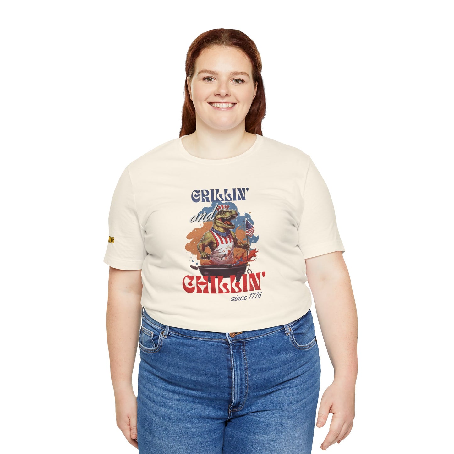 Funny 4th of July BBQ t-shirt featuring a patriotic T-Rex grilling in a red, white, and blue apron, waving an American flag. Perfect for beer and BBQ lovers. Available in plus sizes.