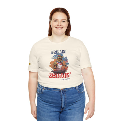 Funny 4th of July BBQ t-shirt featuring a patriotic T-Rex grilling in a red, white, and blue apron, waving an American flag. Perfect for beer and BBQ lovers. Available in plus sizes.