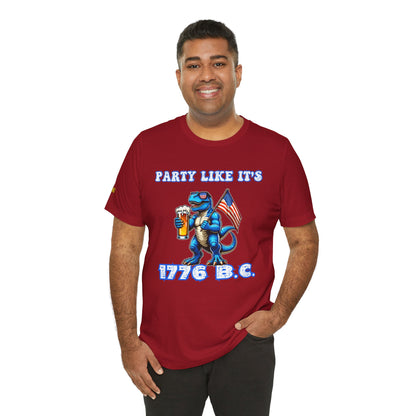 Illustration of a blue T-Rex wearing American flag sunglasses, holding a frosty beer, and waving a USA flag. Funny alcohol quote reads: “Party Like It’s 1776 B.C.” in a bold patriotic font. Canvas red and plus sizes.