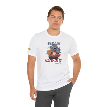 Funny 4th of July BBQ t-shirt featuring a patriotic T-Rex grilling in a red, white, and blue apron, waving an American flag. Perfect for beer and BBQ lovers. In white.
