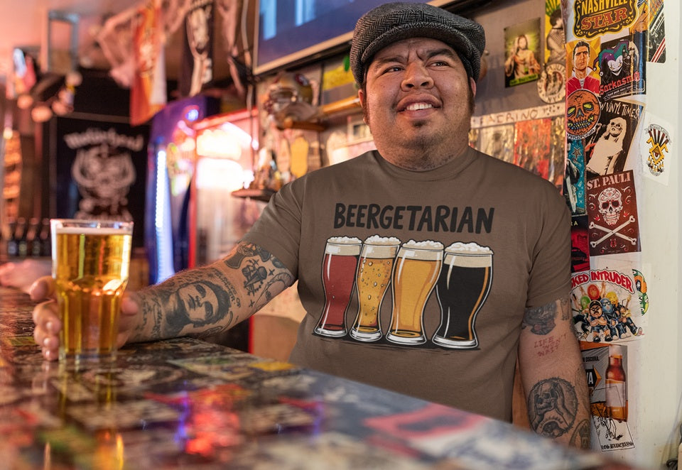 Man with tattoos on arms, sitting against wall in a dive bar holding ice cold beer. Wearing shirt from Beer Strong saying "Beergetarian" across the front, with four large pilsner glasses filled with different beers.